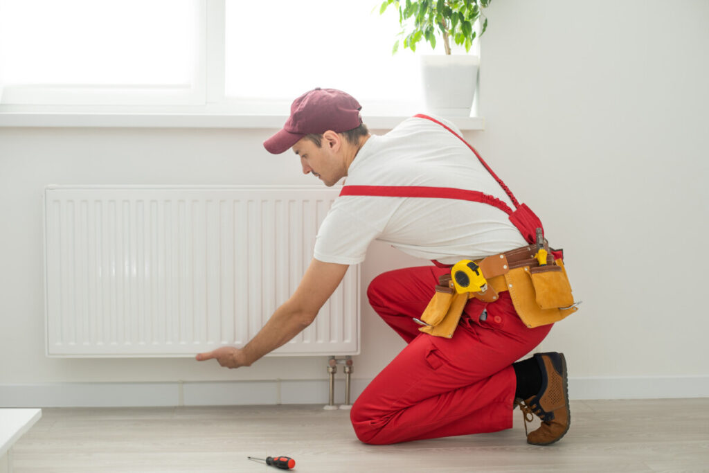 Man in work overalls using wrench while installing heating radiator in room. Young plumber installing heating system in apartment. Concept of radiator installation, plumbing works and home renovation