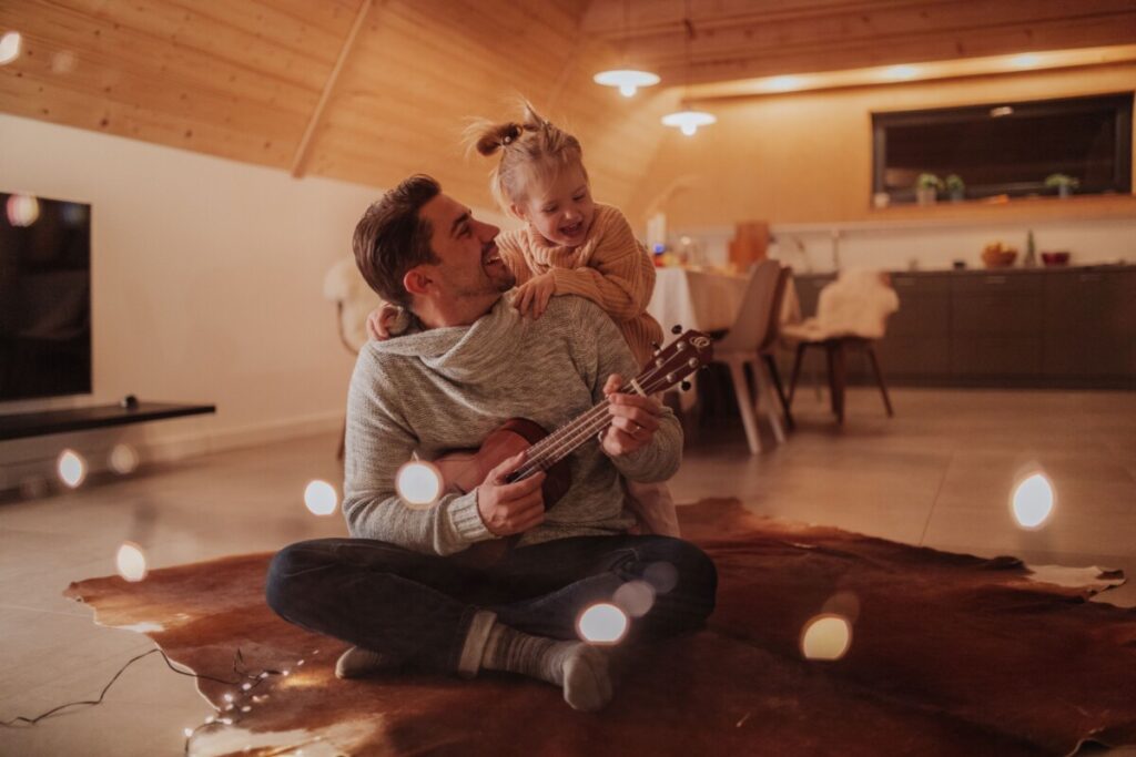 Young cheerful dad sitting with his daughter in living room and playing on guitar.